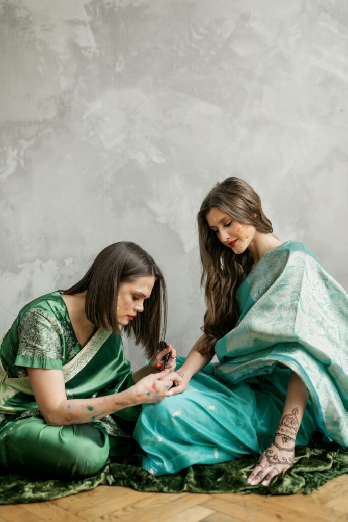 Two women in traditional attire applying mehndi indoors. Vibrant colors and cultural expressions.