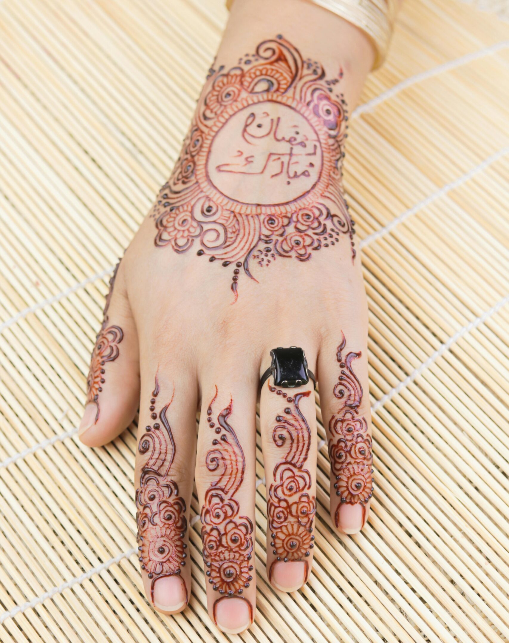 Close-up of a hand adorned with intricate henna art and a black ring on bamboo mat.